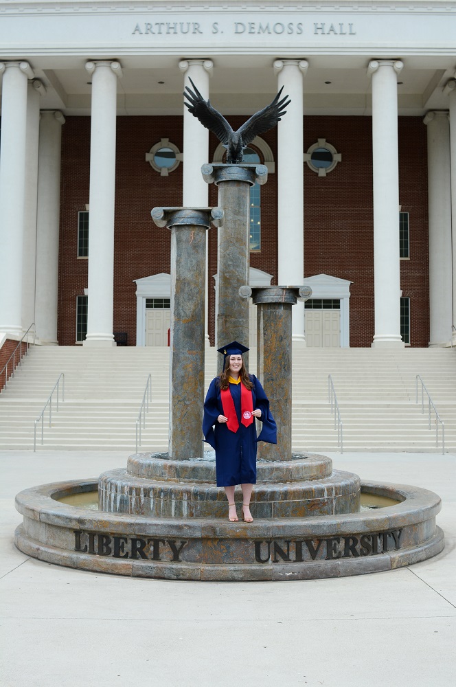 A woman standing in front of the Liberty University eagles in a graduation gown.